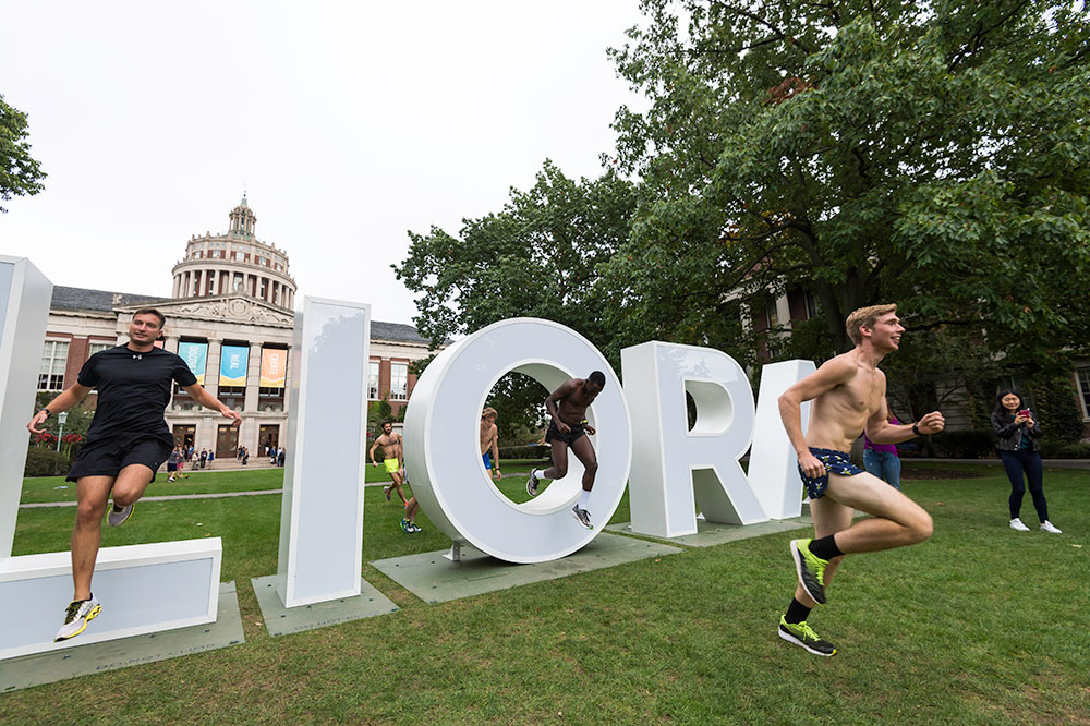 members of the track team running and jumping through large letters that spell MELIORA