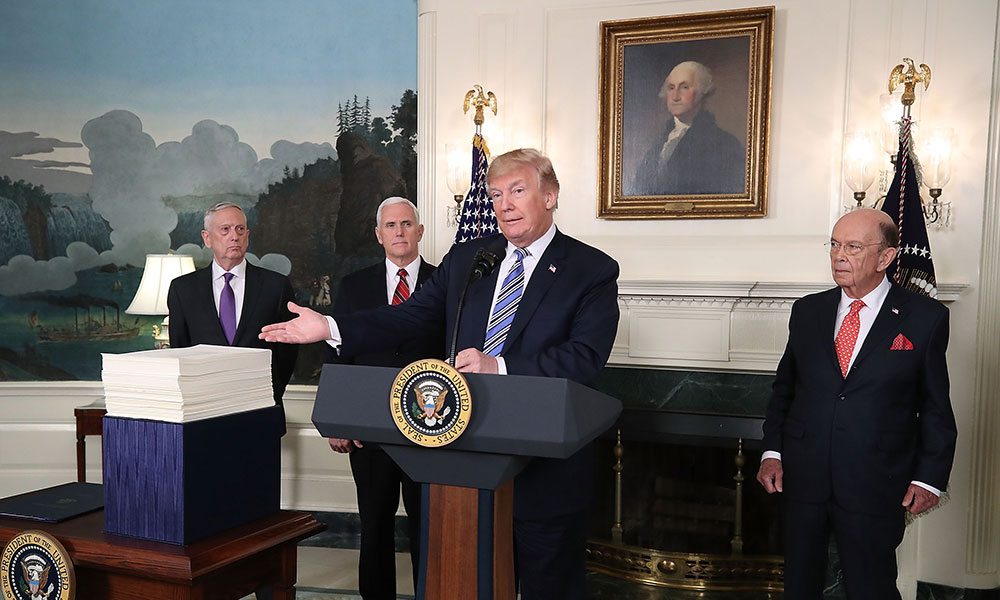 Feature image President Trump points to a large stack of papers, standing behond a podium with three other governement officials