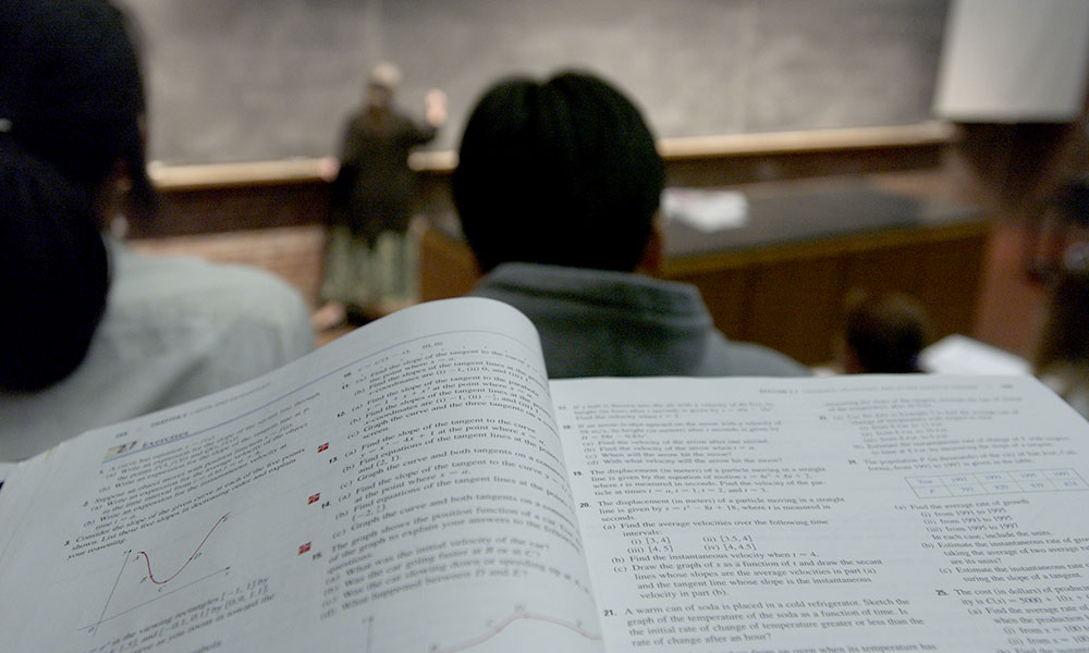 Feature image math text book in front of a student sitting in a large lecture hall