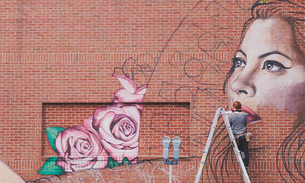 artist on a ladder, painting a mural of a woman with floweres on the side of a building