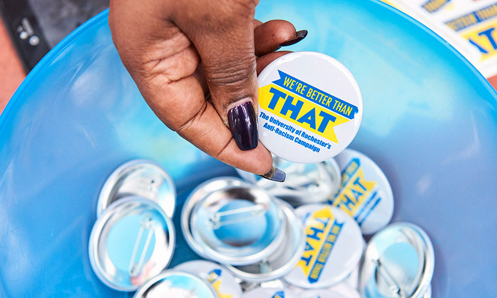 Feature image hand reaches into a bowl of pins that read WE'RE BETTER THAN THAT: THE UNIVERSITY OF ROCHESTER'S ANTI-RACISM CAMPAIGN