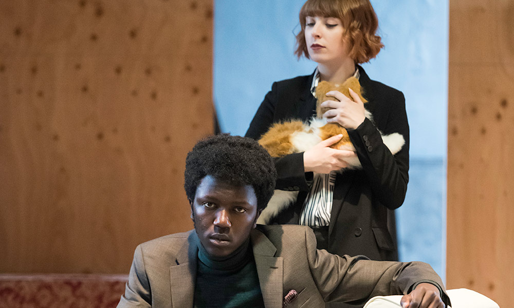 Feature image two actors on stage, one sitting on a couch, the other holding a cat