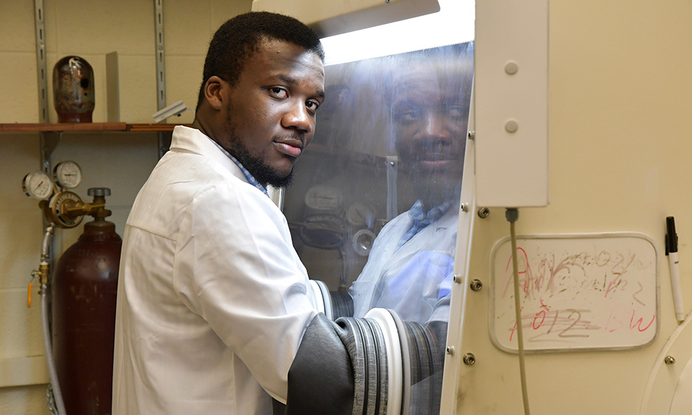 researcher looks into the camera while working with his arms through a glass lab hood