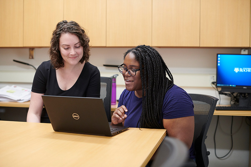 student and professor sitting at a desk, the students is using a laptop to show something to the professor