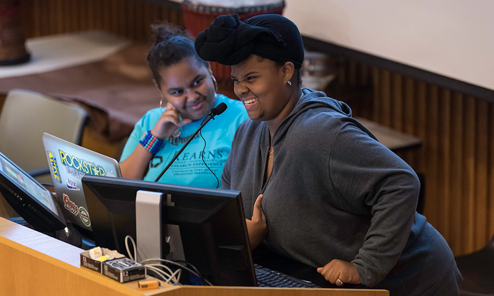 two Upward Bound Rochester students standing at a podium, one with a laptop, one smiling as they speak into the microphone