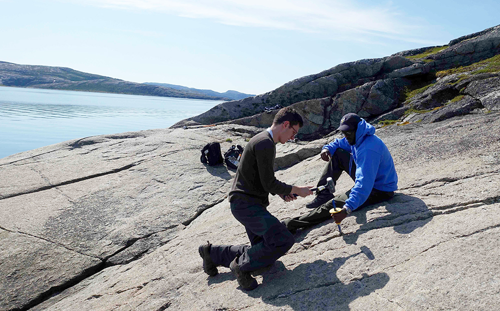 two students on a rock face, using tools to hammer away at the rock surface, with the sun shining on a large body of water in the background