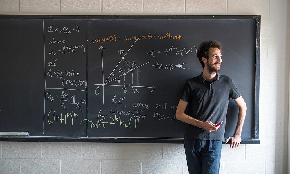 Feature image student smiles while standing in front of a blackboard filled with math symbols
