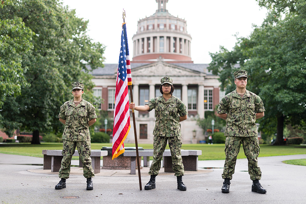 Feature image three Navy midshimpen in uniform stand in front of Rush Rhees Library, the one in the center is holding an American flag