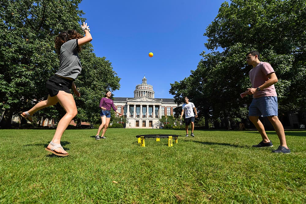 Feature image four students playing a game of spikeball -- hitting a yellow ball into a small round net on the ground -- with Rush Rhees Library in the background