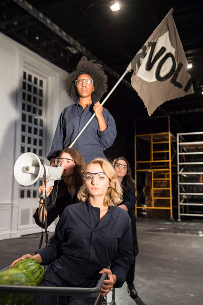 one women pushes three other women in a cart while they wave a flag that reads "Revolt" and use a megaphone