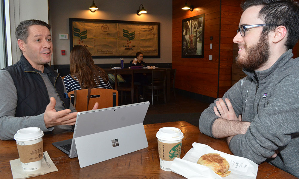 two people sit and talk at a table in a coffee shop, a laptop on the table in front of them