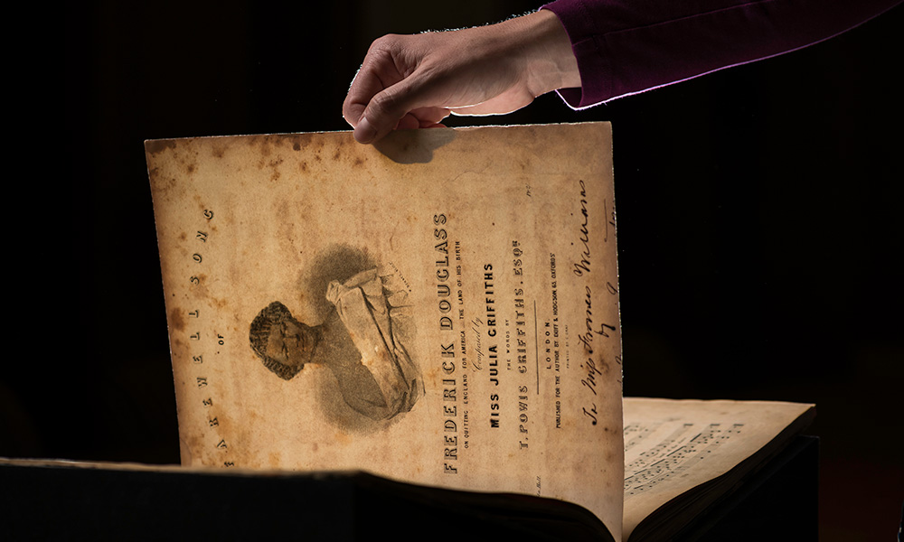 a hand holds open a book of sheet music with an image of Frederick Douglass on the cover and the title FAREWELL SONG FOR FREDERICK DOUGLASS BY MISS JULIA GRIFFITHS