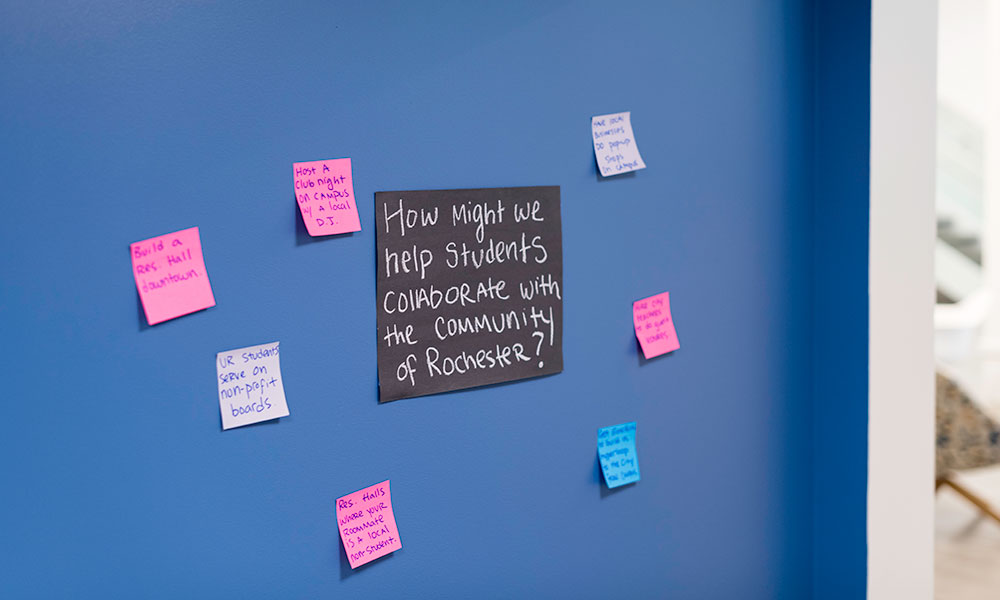 Feature image a blackboard filled with colorful post-it notes, the largest one in the center reads HOW MAY WE HELP STUDENT COLLABORATE WITH THE COMMUNITY OF ROCHESTER