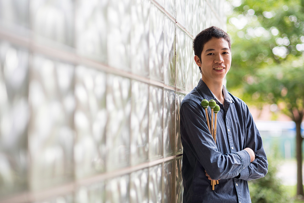 student leans against a glass wall while holding percussion mallets