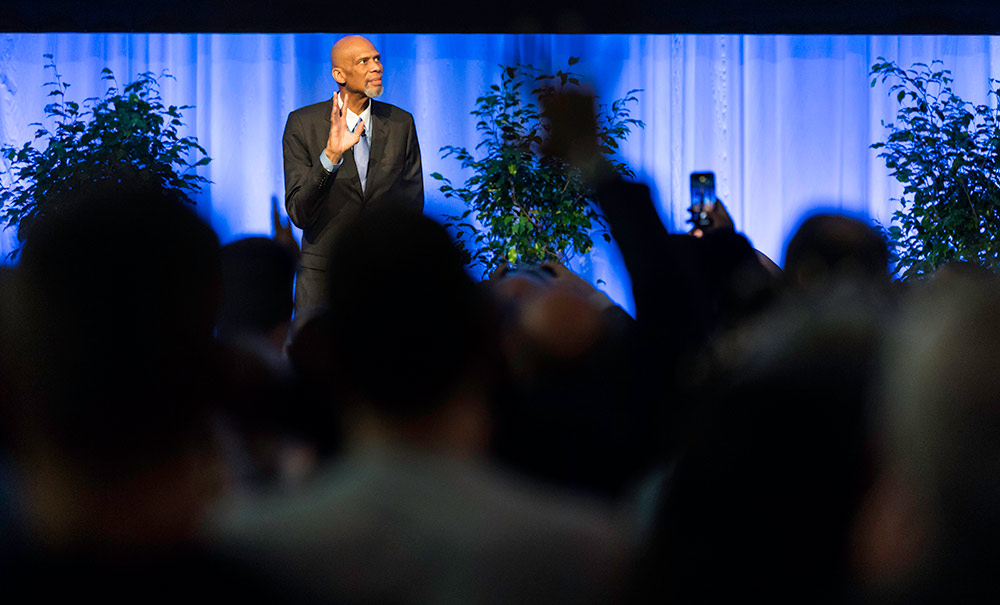 Kareem Abdul-Jabbar stands of stage, towering over the heads of the audience who are also standing in a standing ovatation.