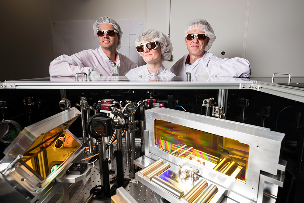 three scientists in clean suits and laser goggles stand in front of an array of optical devices