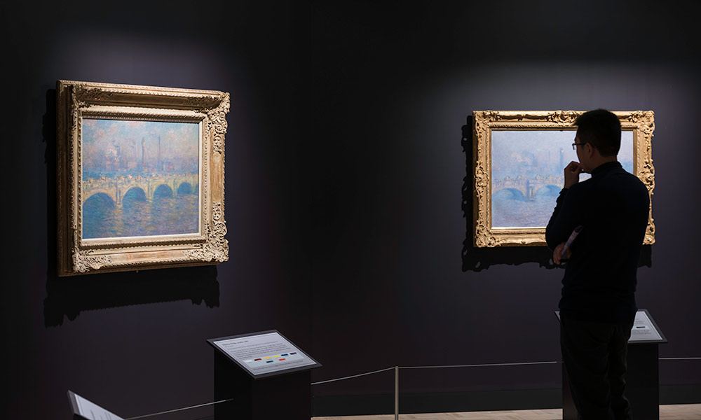 a person in shadow looks at two different Monet paintings, beautifully lit and showcasing different color variations of the same scene.
