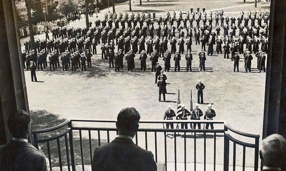 Feature image historical photo of three men on the balcony of Rush Rhees Library looking out over a large assembly of men in uniform on the quad