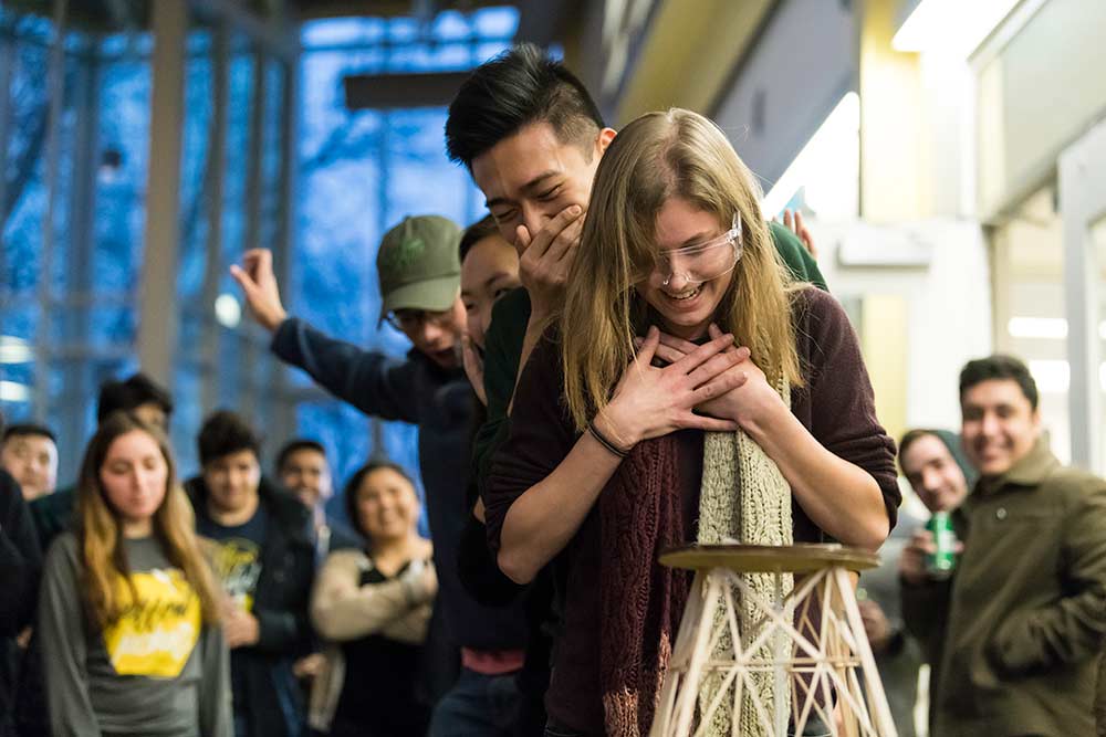 Feature image students gasp and hold their hands over the faces while looking at a fragile tower made of balsa wood slats in the foreground.