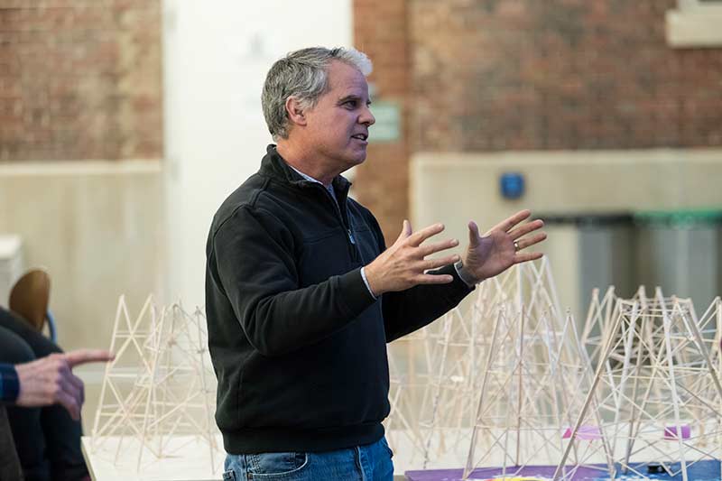 professor speaks to class while standing in front of a large collection of different balsa wood towers.