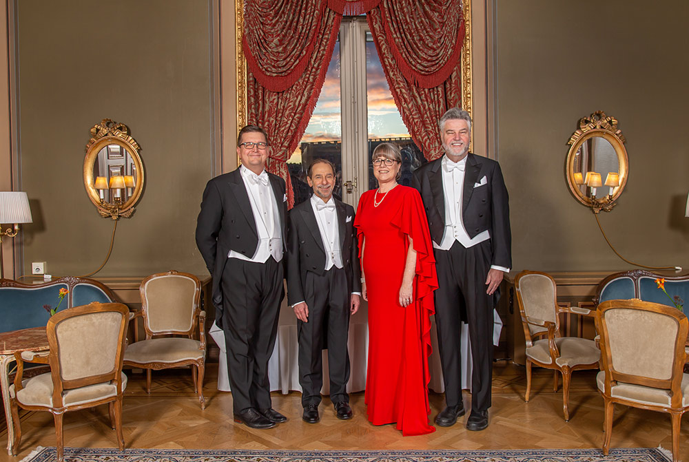 photo of four people in formal attire, white tie and red ball gowns, posing in a grand banquet hall.