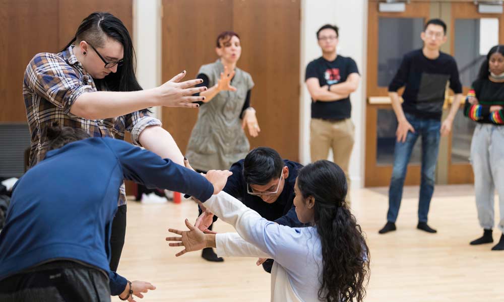 Feature image group of students in the foreground making gestures and movements with hands and arms as another group of students stands in a semicircle behind them
