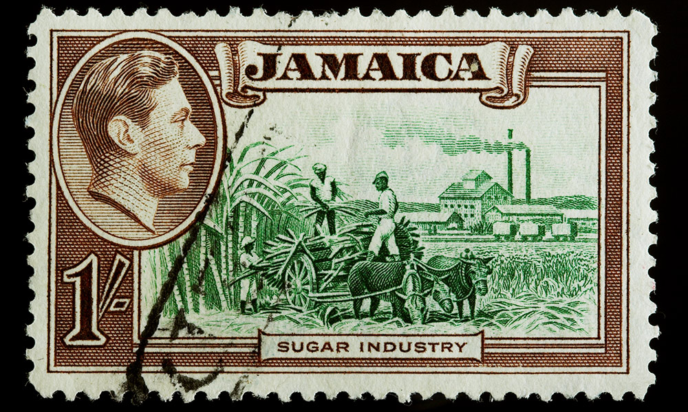 Feature image old stamp from Jamaica includes a photo of King George VI and tow black men working a sugar plantation above the words SUGAR INDUSTRY