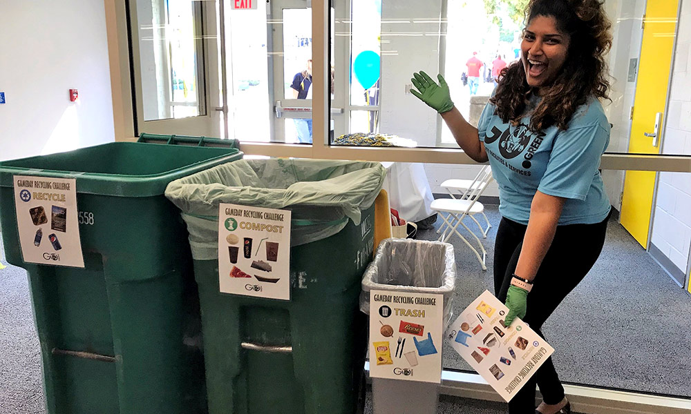 Feature image student in a GO GREEN t-shirt poses next to containers for different types of recycling -- recycle, compost, trash
