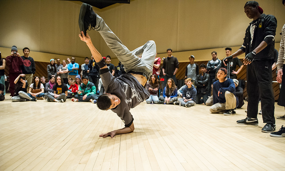 Feature image breakdancer performs a move where he grabs his foot with one hand while standing on the other hand.