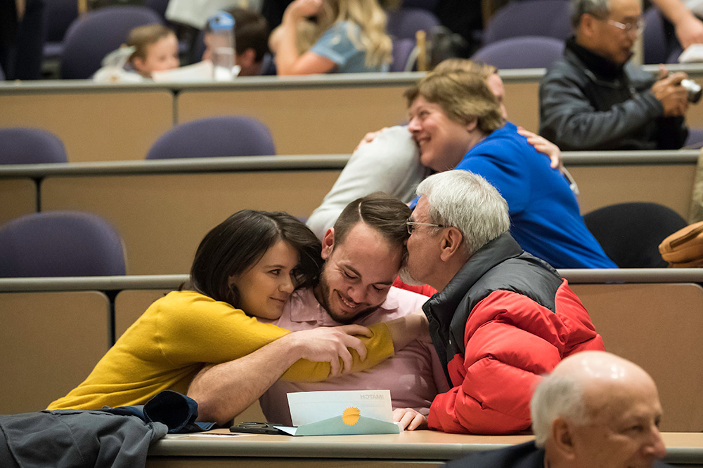 Feature image student holds and envelope while people on either side of him kiss and hug him.