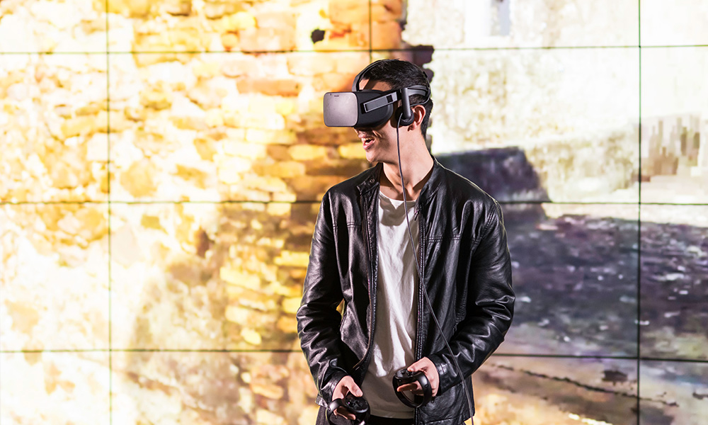 Feature image student wearing virtual reality goggles stands in front of a huge, floor-to-ceiling screen.