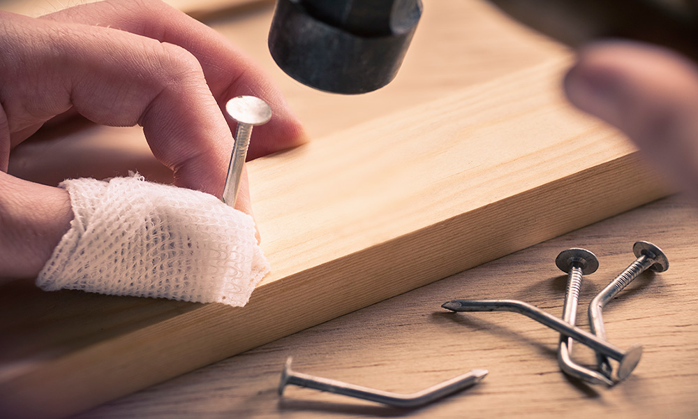 Feature image close up of hands hammering a nail, with a thumb covered in bandages and bent nails all around.