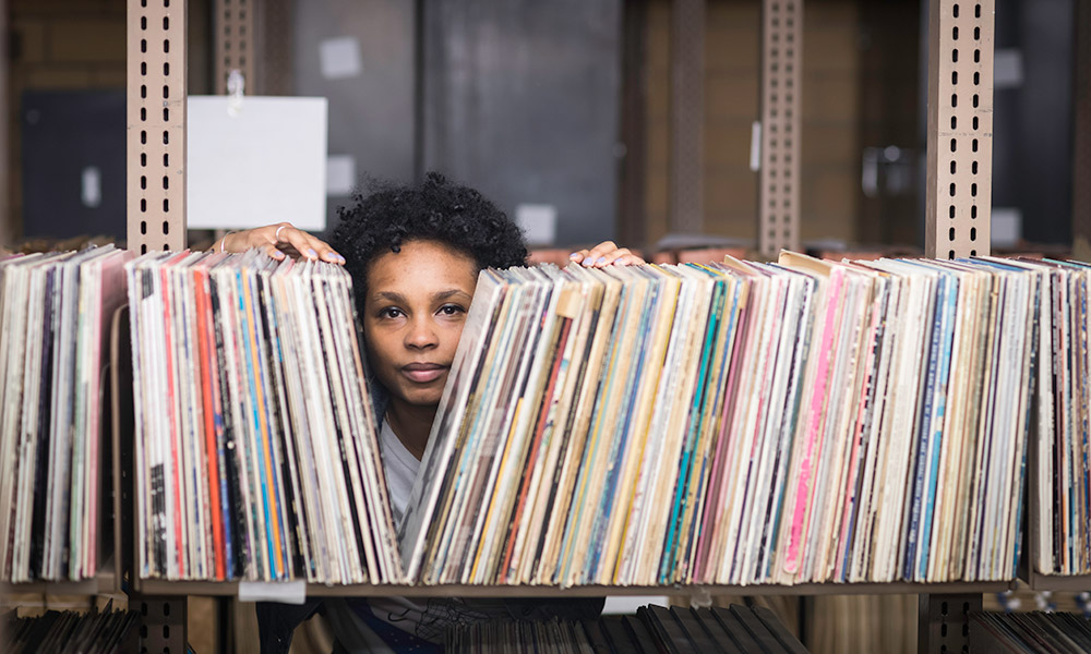 Feature image artist Ash Arder stands in a music library, looking through a long shelf filled with record albums