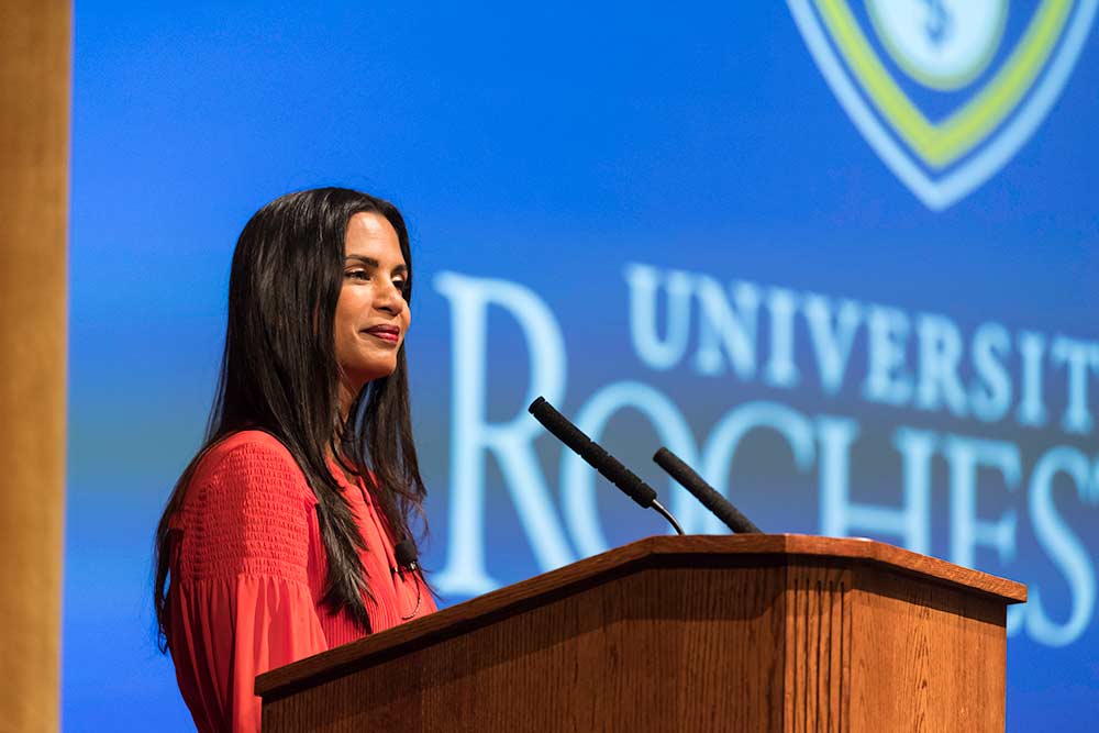 Feature image Speaker stands at a podium on a stage, with the University of Rochester logo projected behind her.