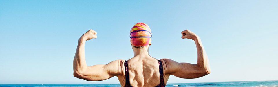 older woman in a swimsuit and cap flexing her muscles at the beach.