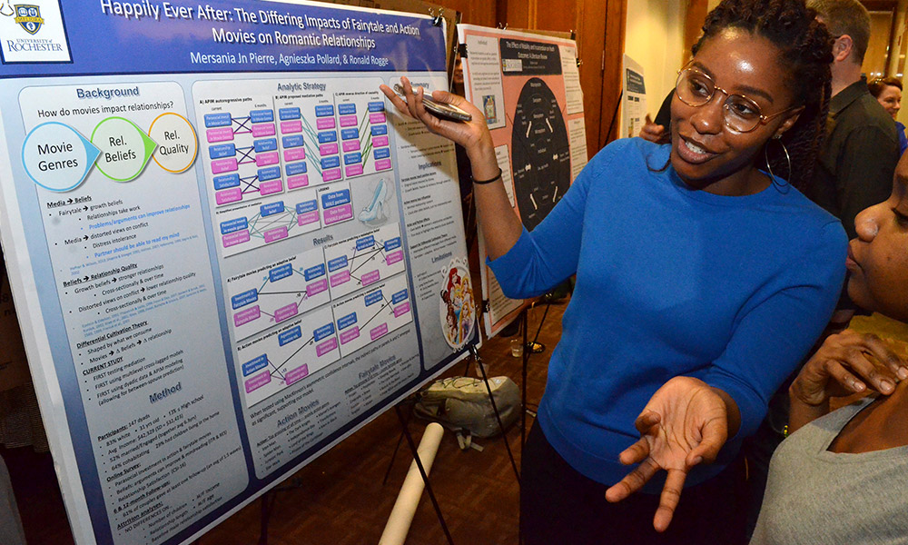 Feature image one student standing in front of a poster,describing her research project to another student.
