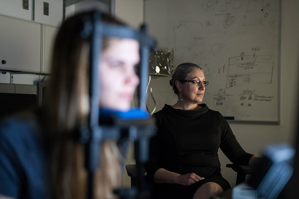 student with her head placed in an eye-examination cradle looks at a screen, while a researcher sits in the background.