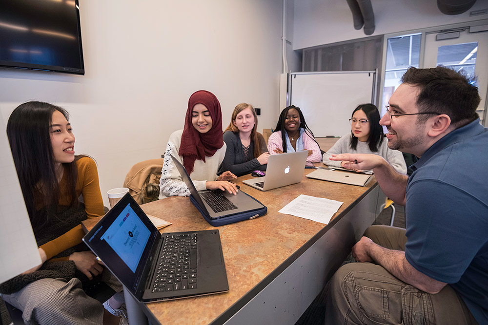 students and professor sit at a long table, each with a laptop in front of them.