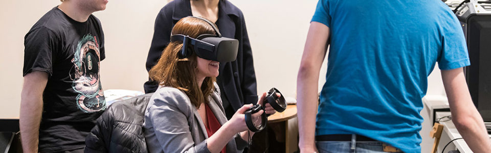 four students -- one in a virtual reality headset -- sitting around a computer screen.