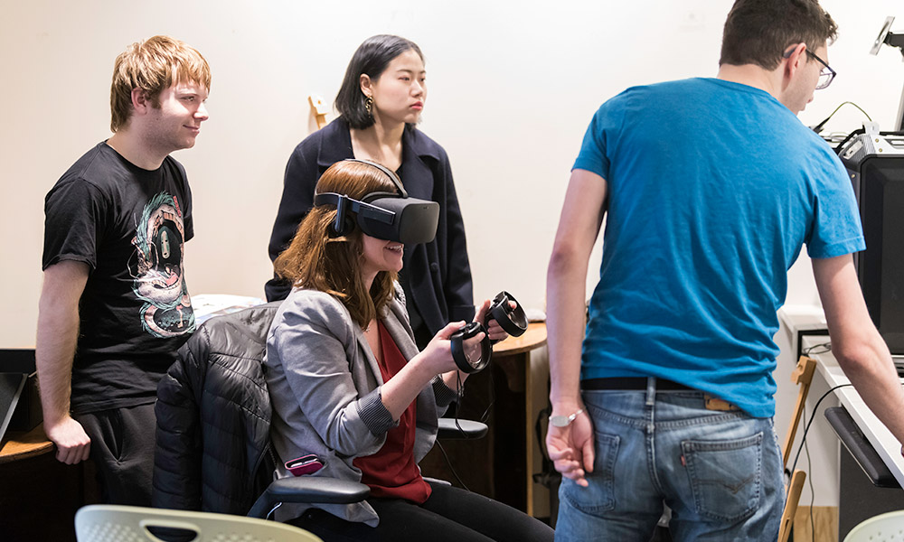 four students, one in a virtual reality headset, sit around a computer screen.