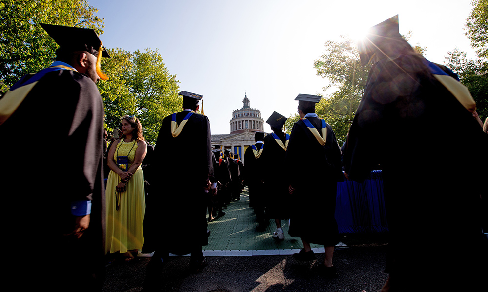 graduates in cap and gown process on the quad toward the library tower.