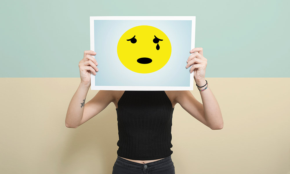 a teenager holds a crying emoticon over their face to symbolize expression of emotions and depression