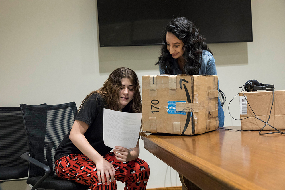 two students reading a script into a microphone and using a cardboard box to muffle the sound. 