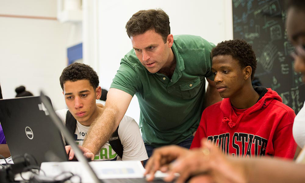 professor leans over the shoulders of two students working on computers