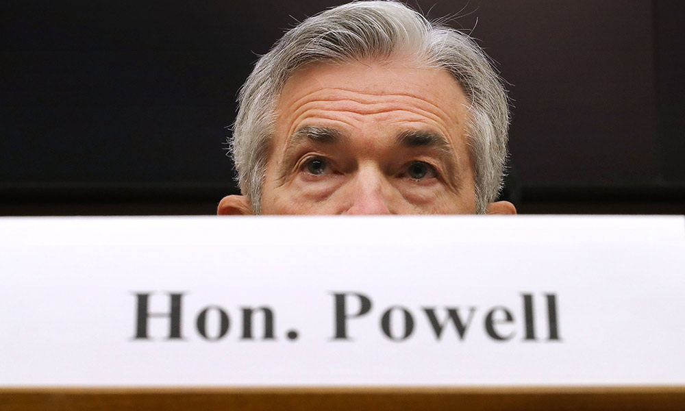 Feature image Jerome Powell sits at a congressional hearing behind a photo of a sign that reads HON. POWELL