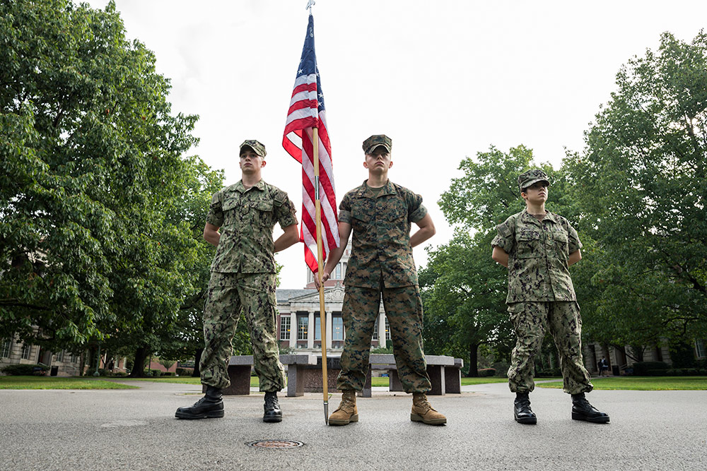 Feature image three navy midshipmen standing and holding an American flag in front of Rush Rhees Library