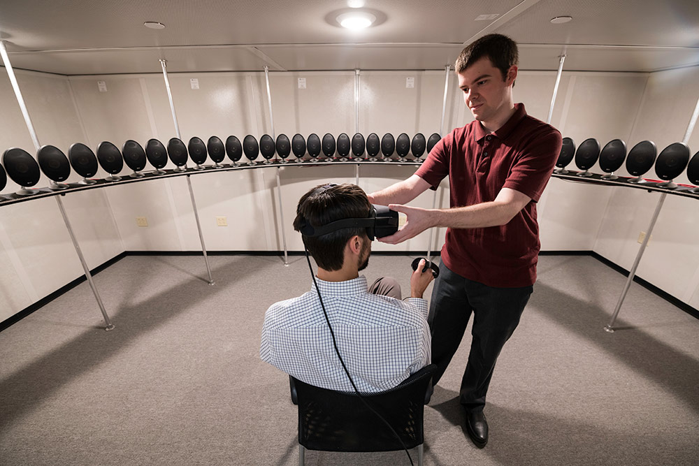 Feature image person sitting in a chair wearing a virtual reality headset and surrounded by speakers as another person adjusts the headset