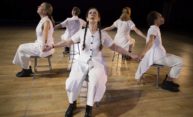 Meredith Monk and four other performers clad in white are seated with their backs to each other while holding hands.