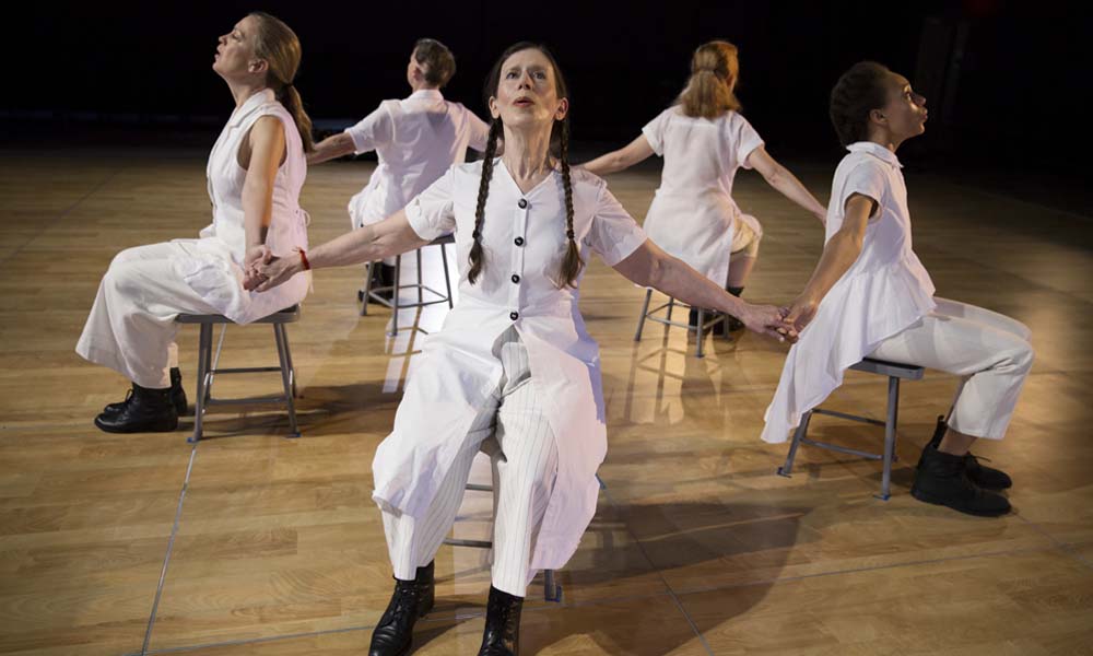 Meredith Monk and four other performers clad in white are seated with their backs to each other while holding hands.