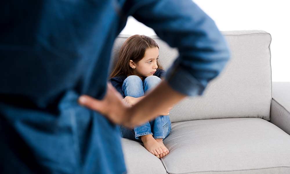 child sitting on a sofa, looking away, mother in the foreground, with her hands angrily on her hips